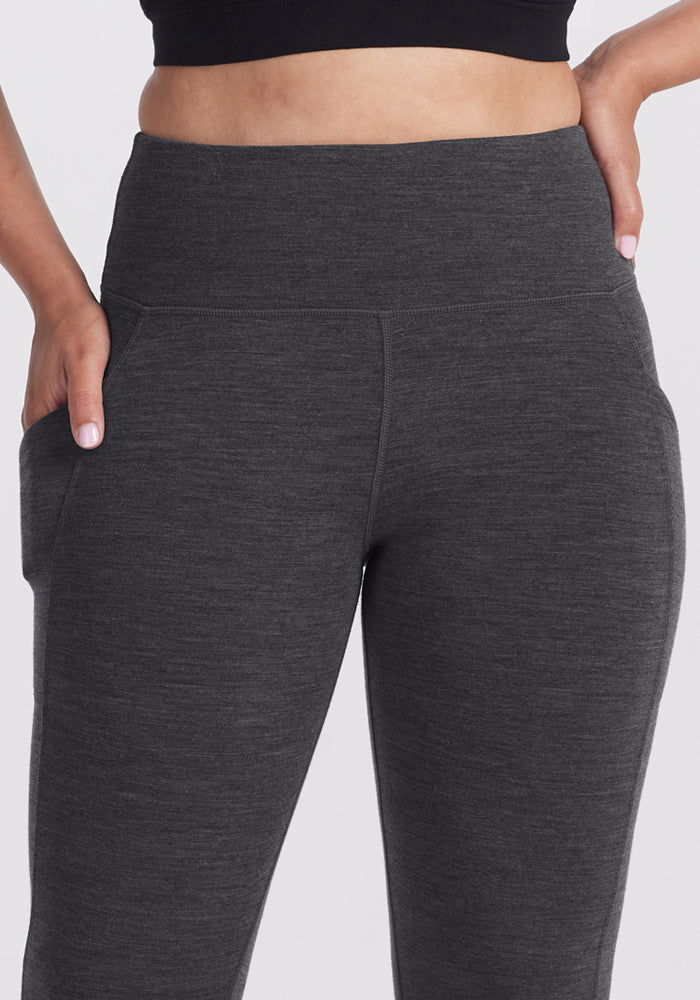 A person wears Woolx Piper Pocket Leggings in Cinder Melange, high-waisted and fitted Merino wool baselayer pants with moisture-wicking comfort, shown from waist to mid-thigh with one hand in the pocket and the other on their hip.