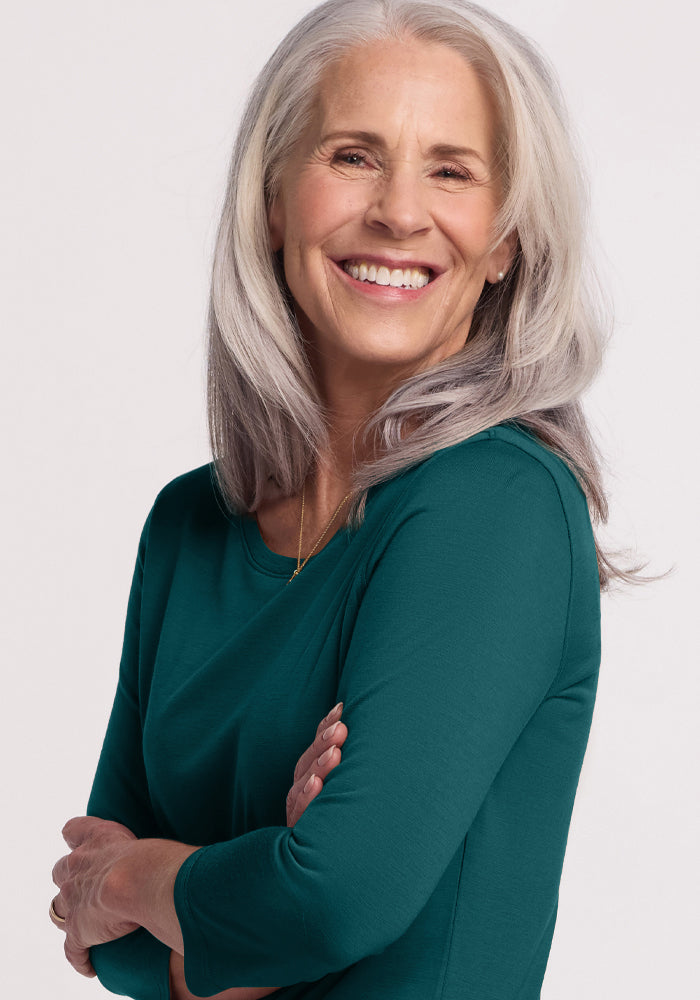 An older woman with long gray hair smiles warmly, arms crossed, wearing the Woolx Jenny 3/4 Sleeve Crew Neck in Riverbank Green, standing against a plain light background.