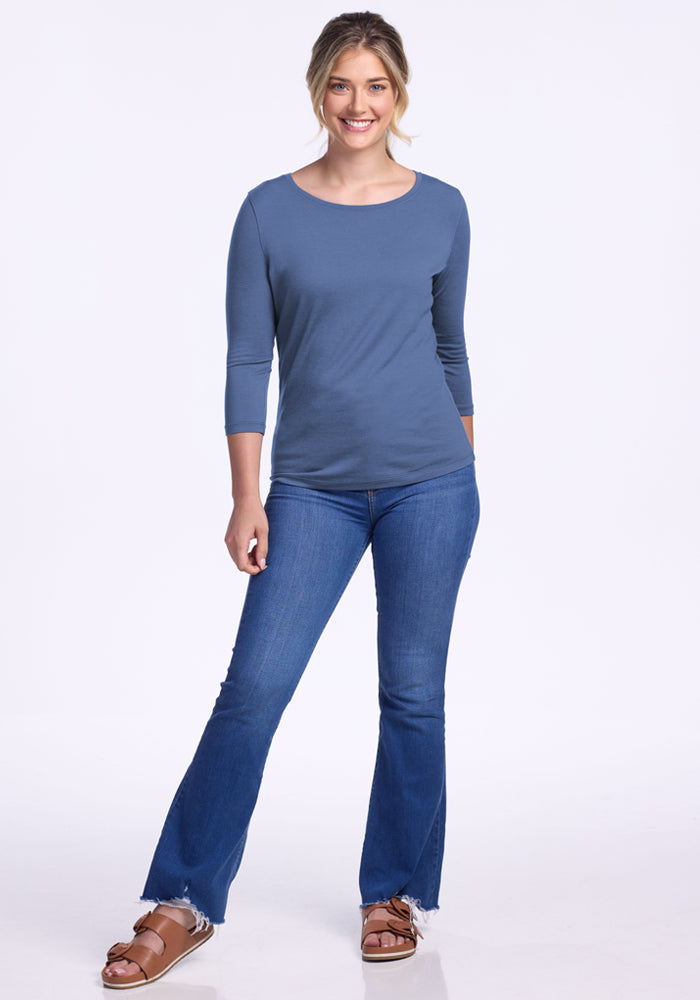 A woman smiles against a white background, wearing the Woolx Jenny 3/4 Sleeve Crew Neck in Skyline, blue jeans with frayed hems, and brown sandals.