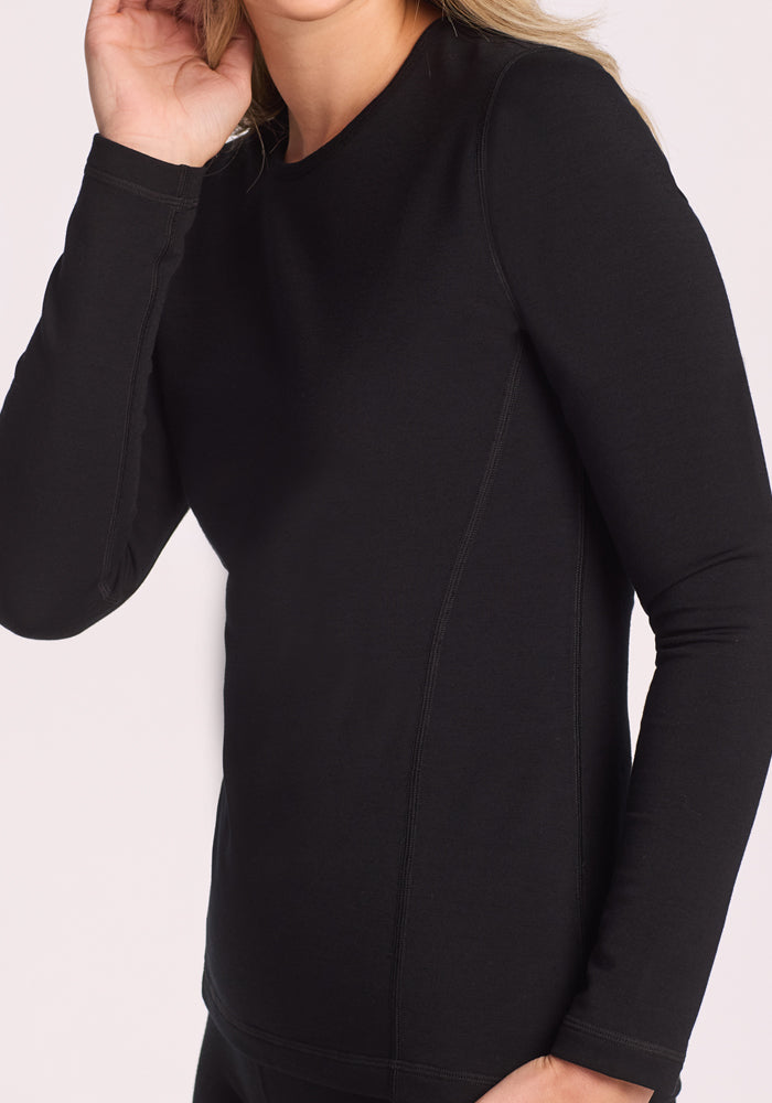 A person models the Woolx Elsa Long Sleeve - Black, a fitted merino wool base layer, standing against a light background with one hand by their side and the other touching their ear.