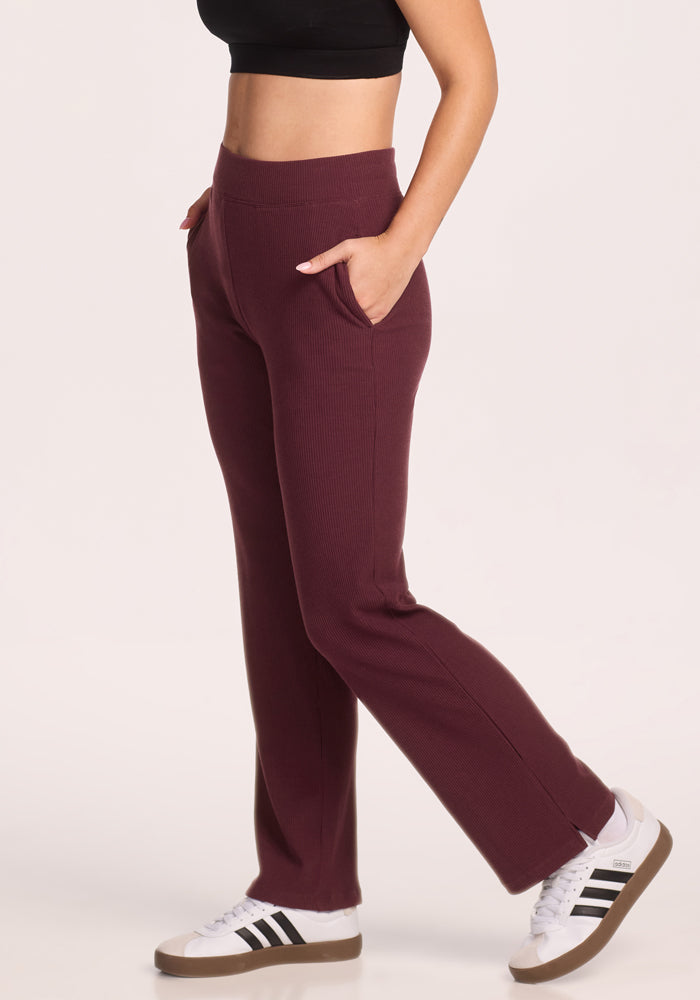 A woman in Woolx Luca Ribbed Pants in Toasted Garnet, paired with a black crop top and white Adidas sneakers with black stripes, stands relaxed against a plain background.