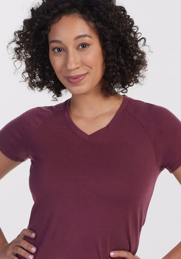 A woman with curly hair wears the Woolx Mia V Neck in Grape Harvest, a short-sleeved wrinkle-resistant travel shirt, and stands smiling with her hands on her hips against a plain light background.