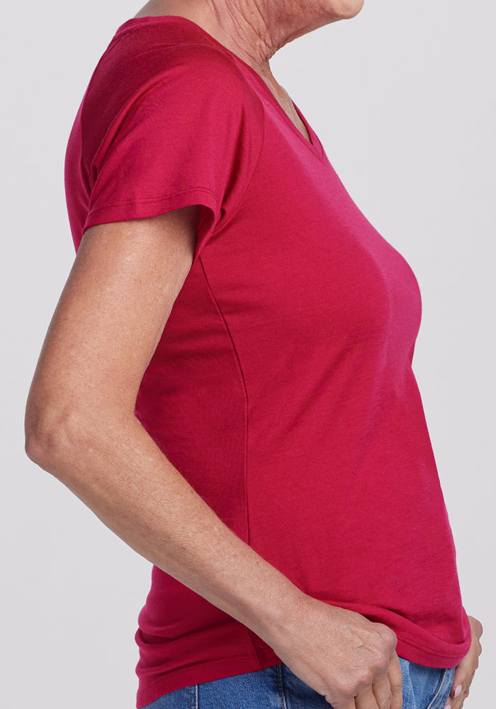 A person is shown from the side against a plain light gray background, wearing the Woolx Mia V Neck - Poppy Spark women's short-sleeved moisture-wicking shirt with blue jeans; only the upper body is visible.