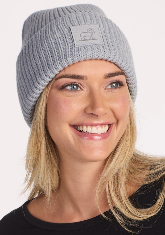 A smiling woman with blonde hair wears the Woolx Ria Chunky Beanie in Morning Fog, a cozy light gray knit winter hat with a front patch, paired with a black top against a plain white background.