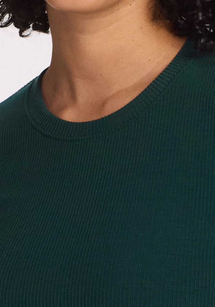 A close-up shows a person wearing the Woolx Hadley Ribbed Crew in Deep Timber. Only the lower half of the face, neck, and upper chest are visible, with curly dark hair partially seen on the right side.