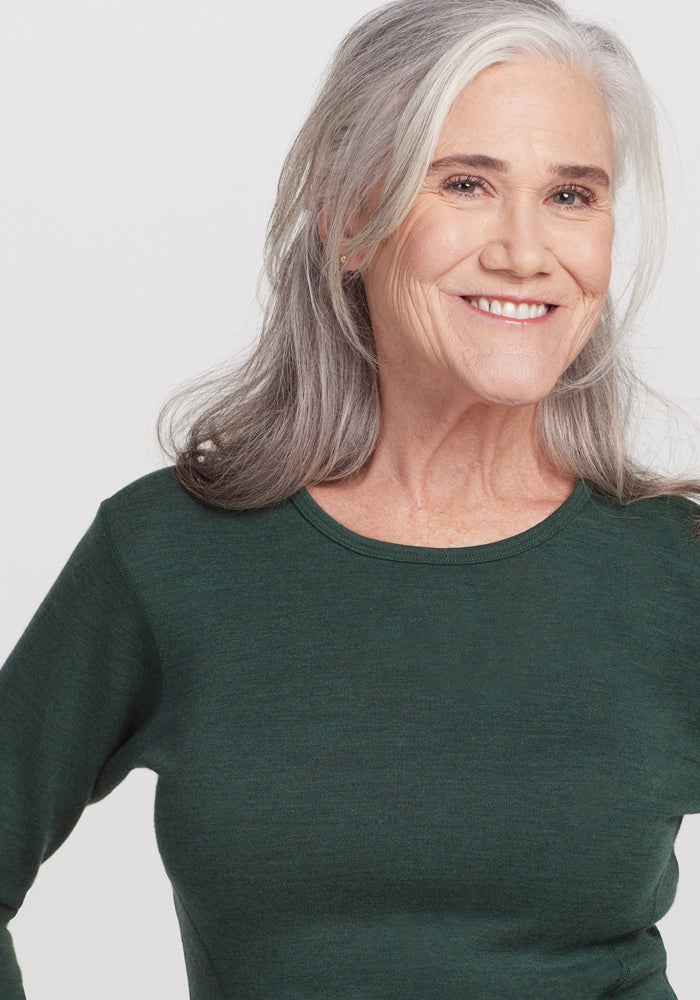 A smiling older woman with long gray hair wears the Woolx Hannah Long Sleeve Top in Forest, a soft, moisture-wicking, odor-resistant shirt, standing against a plain white background.