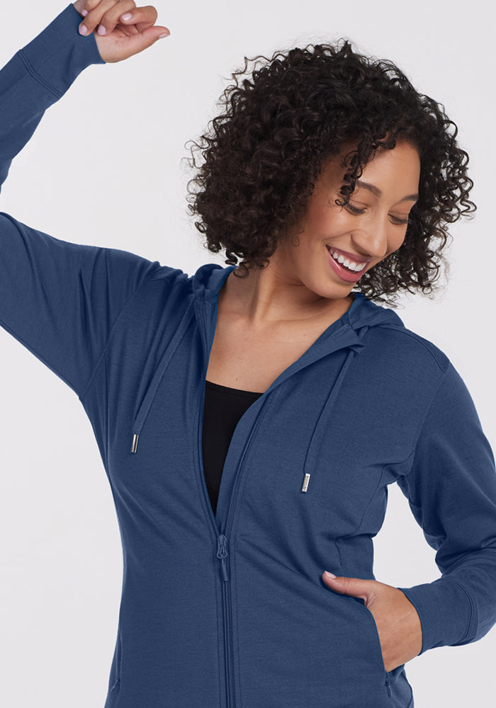 A woman with curly hair smiles while raising one arm, wearing the Woolx Ryann Hoodie - Starry Night over a black top, with her other hand in her pocket against a light background.