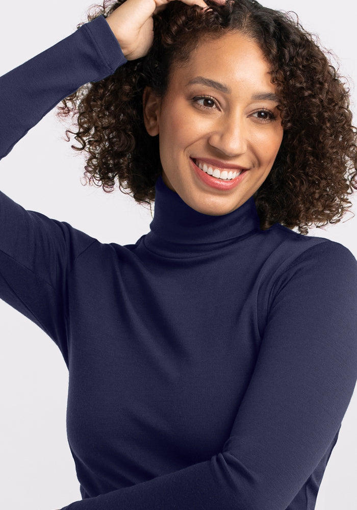 A woman with curly hair smiles while looking to the side, wearing the Woolx Peyton Turtleneck in Midnight Sapphire. She raises one hand to touch her hair against a plain, light-colored background.