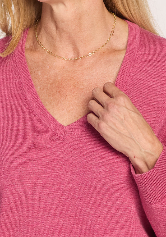 A person wearing the Woolx Francesca Sweater in Wild Salmon and a delicate gold chain necklace touches their collarbone with one hand against a plain background.