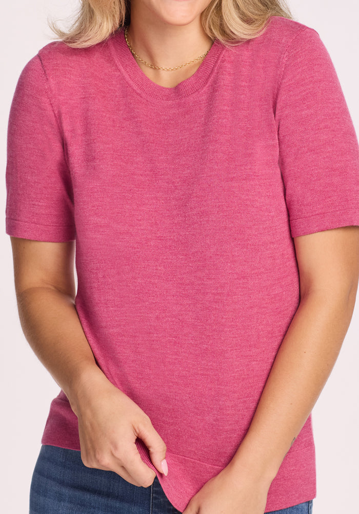 A woman with shoulder-length blonde hair wears the Woolx Anna Crew Sweater in Wild Salmon and blue jeans, holding the hem of her shirt. Her face is partially out of frame.