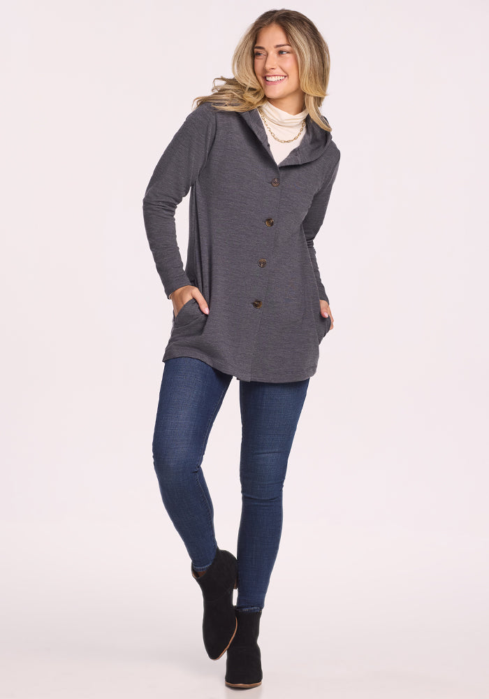 A smiling woman with long blonde hair wears the Woolx Edna Hooded Cardigan in Cinder Melange, paired with blue skinny jeans and black ankle boots. She stands against a plain light background, hands in her pockets—perfect for chilly days.