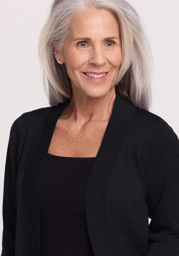 A smiling older woman with long gray hair models the Woolx Macie Sweater in black, paired with a black top, standing against a plain light background.