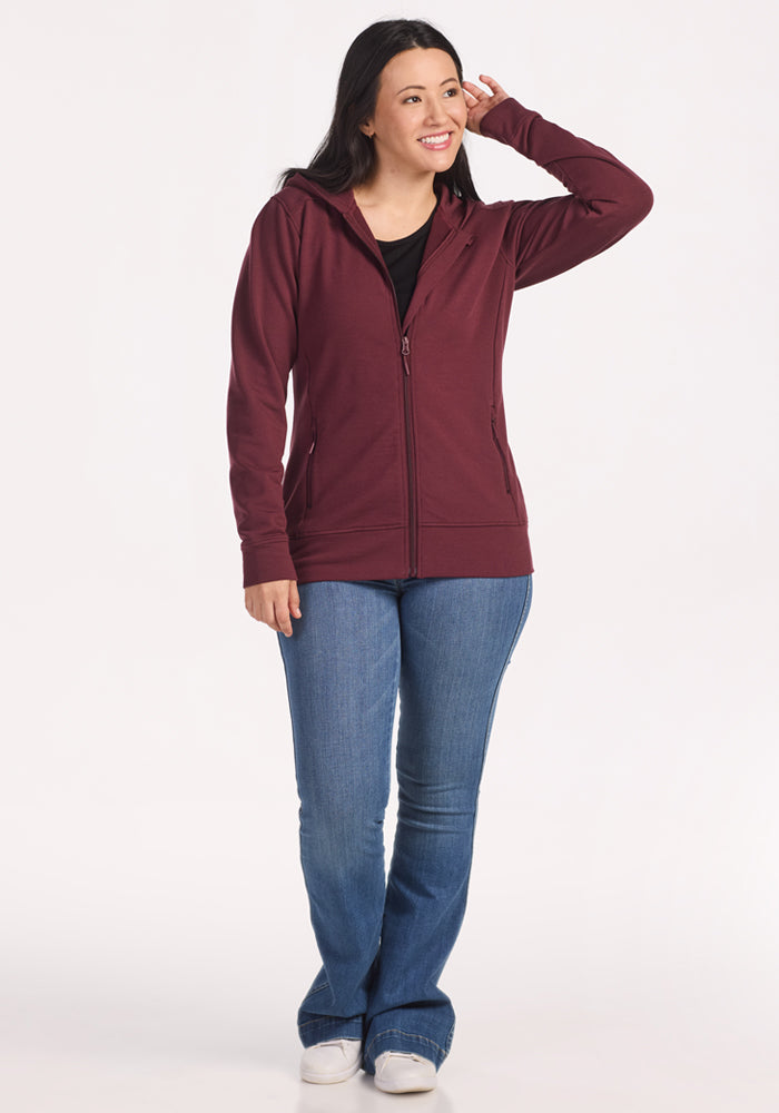 A smiling woman with long dark hair stands against a white background, wearing the Woolx Darby Zip Up Hoodie in Toasted Garnet, paired with blue jeans and white sneakers. She touches the side of her head with one hand.