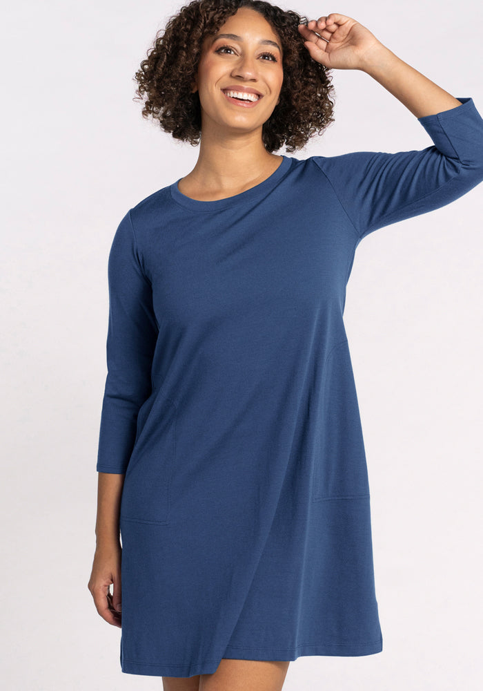 A woman with curly hair smiles in the Woolx Rissa A Line Dress - Starry Night, a blue, knee-length, long-sleeved merino wool dress—ideal for fall and winter layering. One hand is near her head, the other at her side, against a light background.