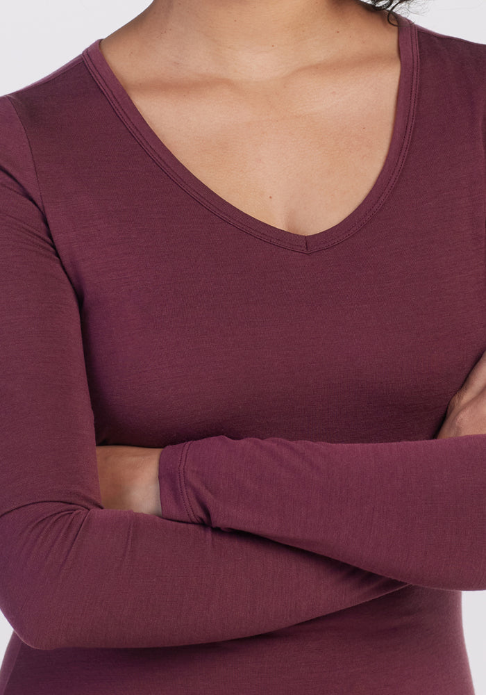 A woman with arms crossed is shown from neck to waist wearing the Woolx Layla V Neck Top in Grape Harvest, a long-sleeve travel shirt.