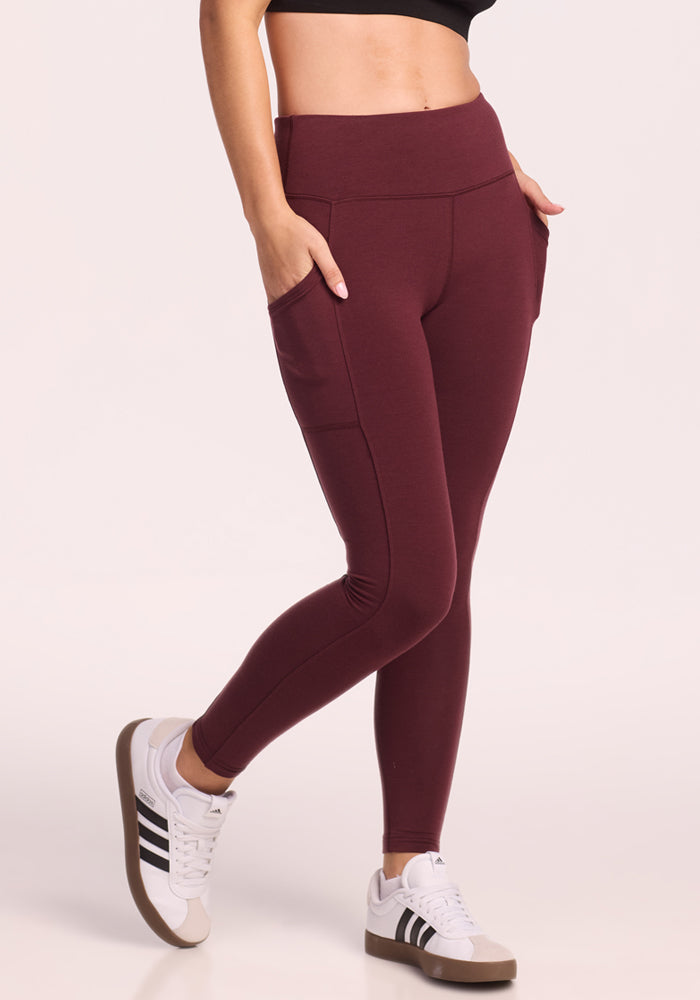 A woman models Woolx's Piper Pocket Petite leggings in Toasted Garnet, featuring high-waisted design and side pockets, paired with a black crop top and white sneakers with black stripes, standing casually against a light background.