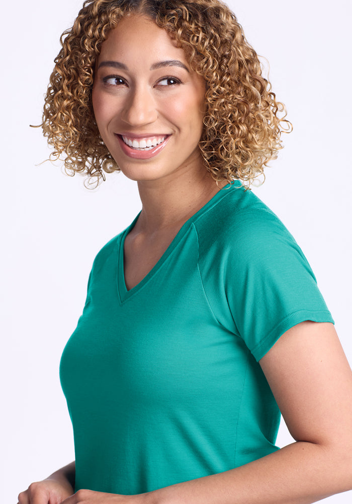 A woman with curly, shoulder-length hair smiles while wearing the Woolx Mia V Neck - Cool Jade, looking slightly to the side against a plain background, highlighting this Merino wool tee’s comfort and moisture-wicking benefits.