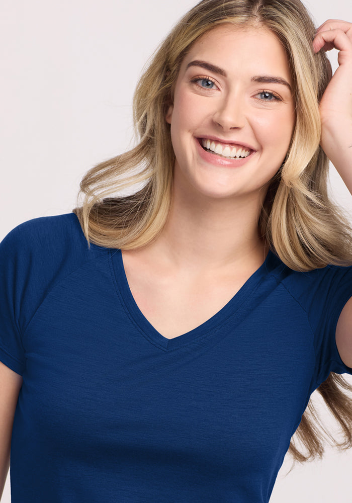 A smiling young woman with long blonde hair wears the Woolx Mia V Neck in Estate Blue, posing with one hand touching her hair against a light background.