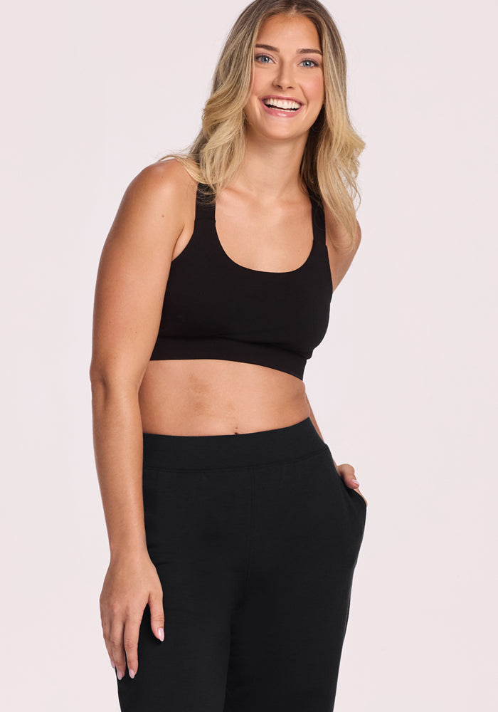 A smiling woman with long blonde hair wears a black sports bra and Woolx Brynn Petite Pants in black, standing with one hand in her pocket against a plain light background.