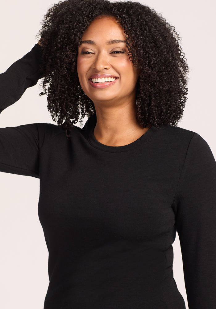A woman with curly hair smiles, raising one hand to her head, while wearing the Woolx Sadie Baselayer Top - Black against a light background.