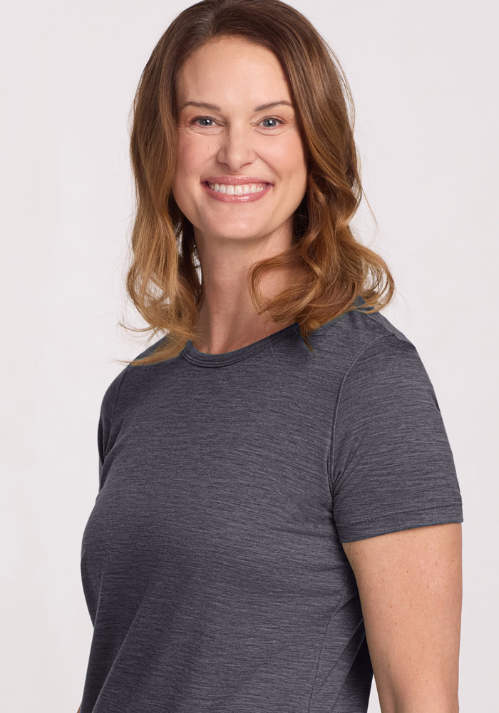 A woman with wavy, shoulder-length light brown hair smiles while wearing the Woolx Liza Crew Neck Tee in Cinder Melange. She is shown from the waist up against a plain, light background.
