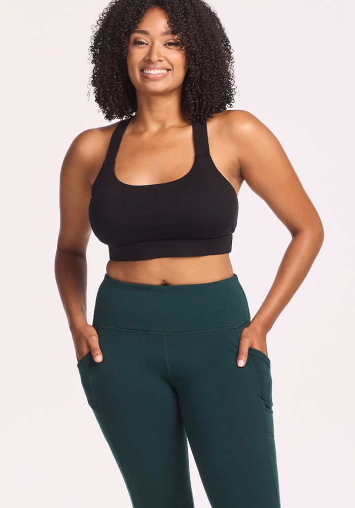 A woman with curly hair smiles in Woolx’s Piper Pocket Petite leggings in Deep Timber and a black sports bra, standing against a plain light background. The leggings, designed for petite women, have pockets and are perfect for cold weather.