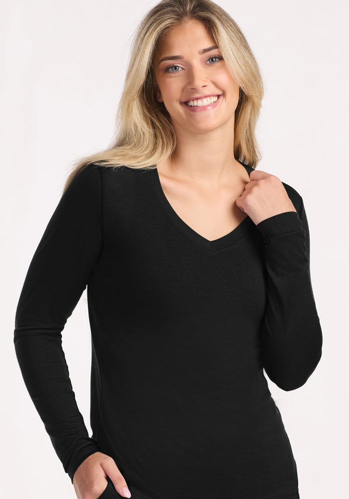 A smiling woman with long blonde hair wears the Woolx Lily V Neck Lounge Top in black, a fitted long-sleeve sleep shirt, as she stands against a plain light background.