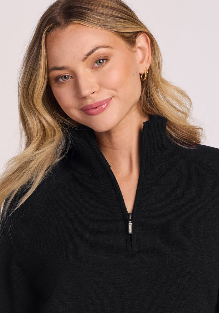 A young woman with wavy blonde hair smiles gently, wearing the Woolx Collins Half Zip Sweater in black and gold hoop earrings, posed against a plain light background.