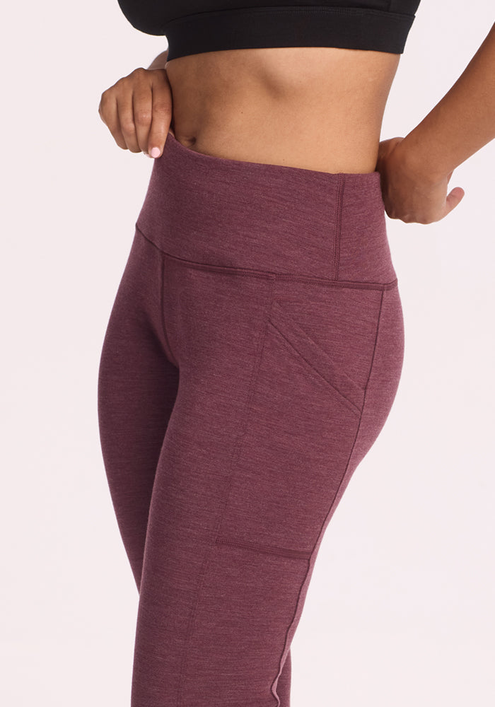 A person wearing Woolx Frost Duralite® Leggings in Grape Harvest Melange, shown from mid-torso to thigh, adjusts the high waistband; the leggings feature side pockets.