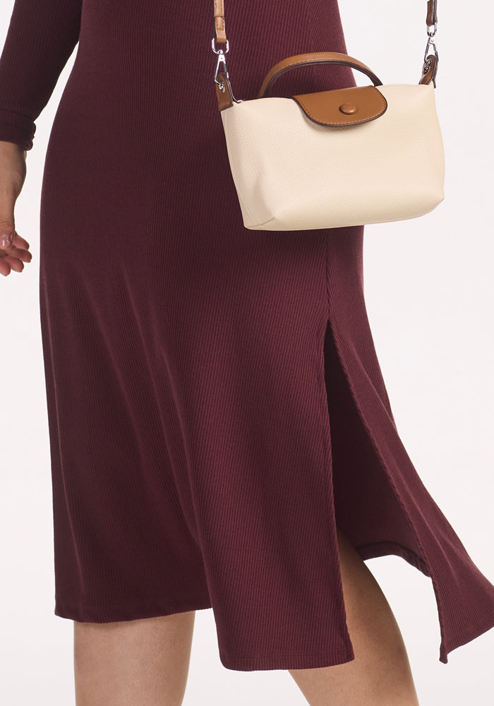 A person is wearing the Woolx Caroline Ribbed Twirl Dress in Toasted Garnet, featuring a side slit, and holding a small beige handbag with brown leather accents and a shoulder strap.