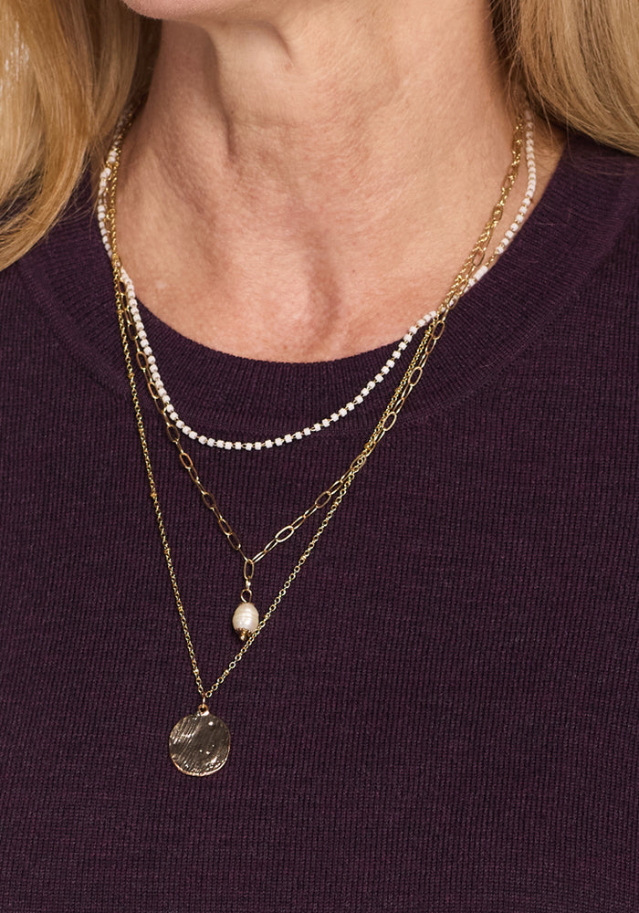 A close-up of a person wearing the Woolx Anna Crew Sweater in Deep Plum, styled with three layered necklaces: one featuring small white beads, another with a single pearl, and a third with a round textured gold pendant.