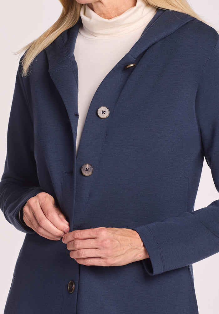 A person fastens a button on the Woolx Edna Hooded Cardigan in Deep Navy, worn over a white turtleneck. The photo highlights the upper body, hands, and cozy merino wool—ideal for cold-weather comfort.