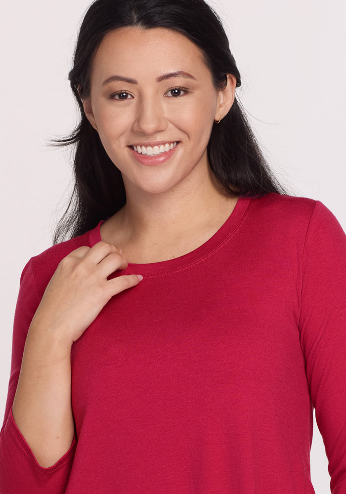 A woman with long dark hair smiles at the camera, wearing the Woolx Rissa A Line Dress in Poppy Spark—a bright pink Merino wool dress perfect for fall and winter. She gently touches her collar against a plain light background.
