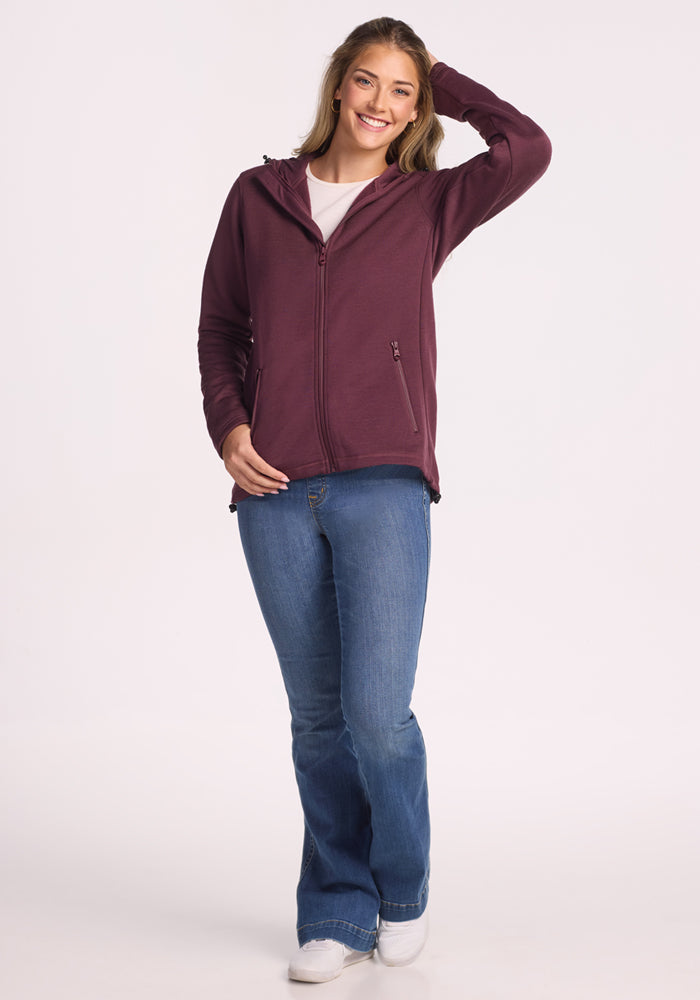 A woman stands smiling with one hand on her head, wearing the Woolx Cubby Hooded Sweatshirt in Grape Harvest, blue jeans, and white shoes against a plain white background.