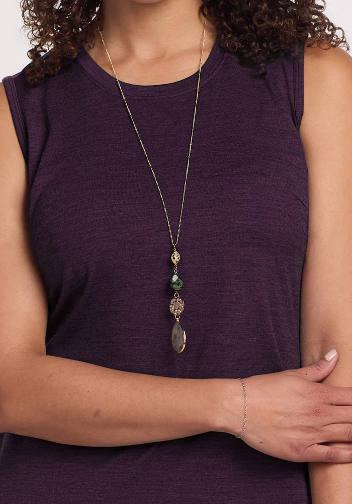 A woman in the Woolx Cassie Dress in Deep Plum wears a long gold necklace with beads and a teardrop pendant, her curly hair partially visible and her left arm resting across her body.
