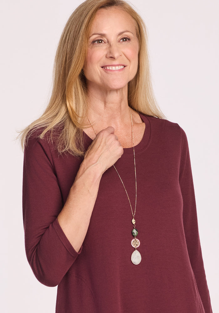 A smiling woman with long blonde hair wears the Woolx Rissa A Line Dress in Toasted Garnet and a layered necklace, perfect for fall and winter, standing against a plain light background.
