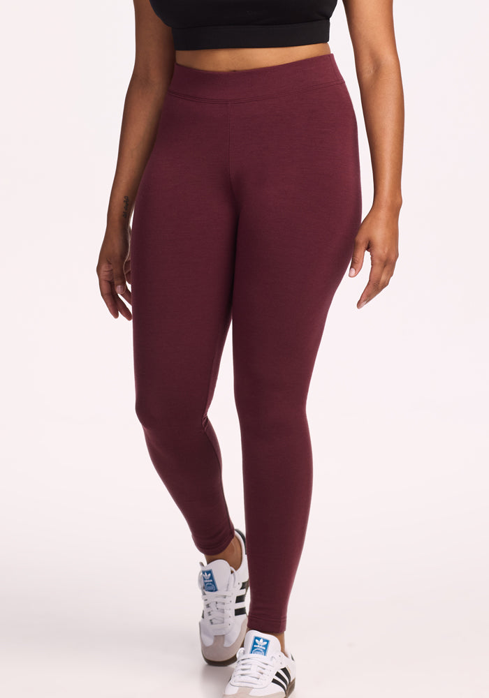 A person wearing a black crop top, Woolx Stella Leggings in Toasted Garnet, and white Adidas sneakers with blue logos stands against a plain white background. Only the lower half of their body is visible. 