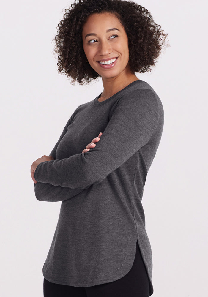 A woman with curly hair smiles, arms crossed, wearing the Woolx Blair Tunic in Cinder Melange—offering lightweight warmth and style against a plain light background.