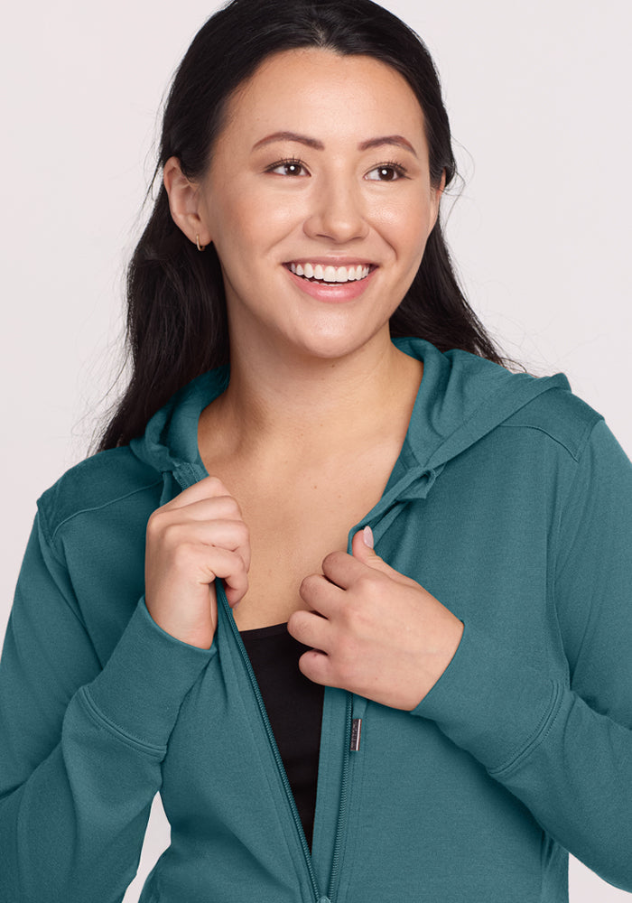 A woman with long dark hair smiles as she zips up the Woolx Ryann Hoodie in Tidepool over a black top against a light background—an ideal travel essential.