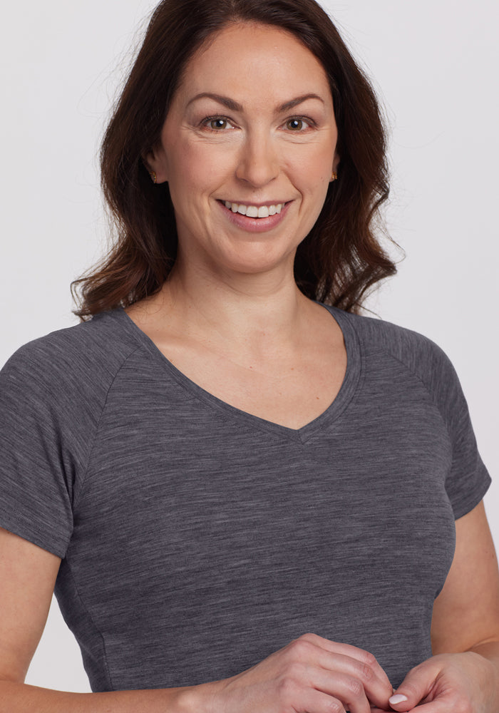 A woman with wavy brown hair, wearing the Woolx Mia V Neck - Cinder Melange tee, smiles while standing against a plain light background.
