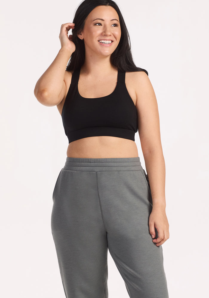 A woman with long dark hair smiles while wearing a black crop tank and Woolx Parker Sweatpants in Sedona Sage. She stands against a plain light background, one hand touching her hair and the other by her side.