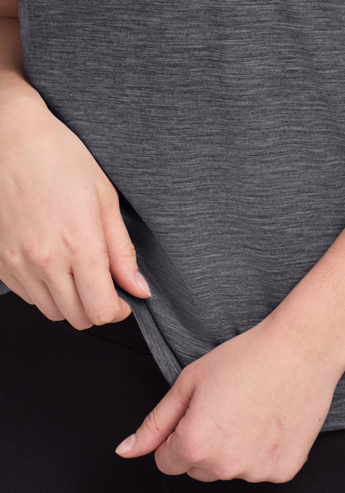 Close-up of hands adjusting the hem of a Woolx Junie V Neck tee in Cinder Melange, a women’s relaxed fit Merino wool top with moisture-wicking properties. The person is wearing black pants against an indistinct background.