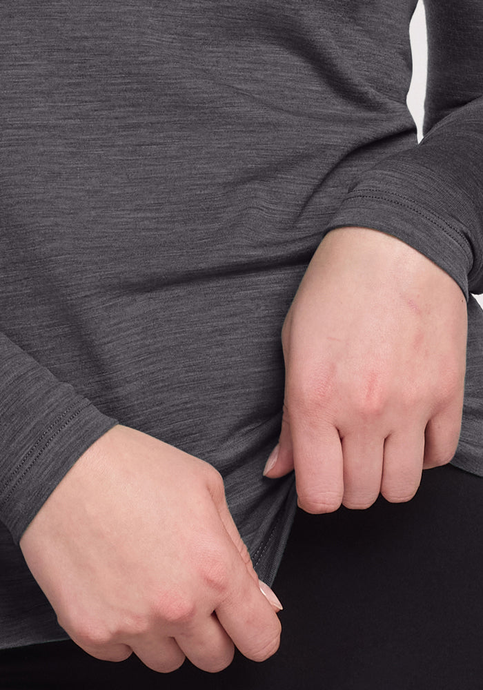 Close-up of hands holding the hem of a gray Woolx Remi Long Sleeve T-Shirt in Cinder Melange, paired with black pants. Image highlights fabric texture and moisture-wicking properties.
