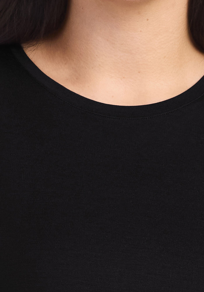 A close-up of a person wearing the Woolx Billie Lounge Tee in black, featuring a plain crew neck that reveals part of the neck, collarbone, and upper chest. The background and lower face are not visible.