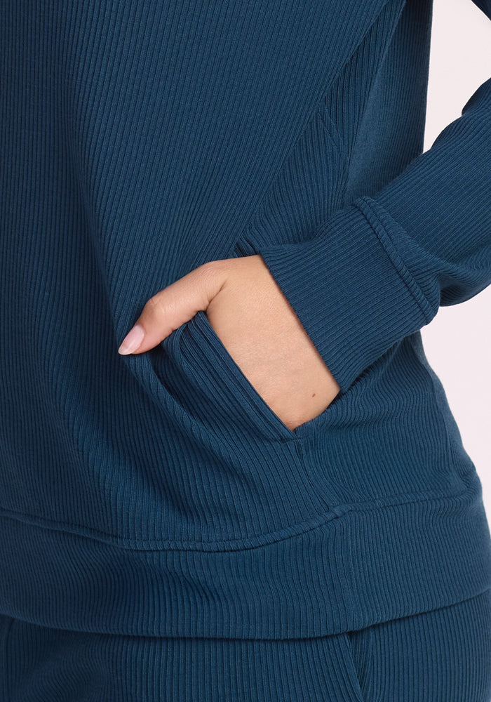 A person’s hand is partially in the pocket of the Woolx Quinn Ribbed 1/4 Zip in Majolica Blue, paired with matching ribbed blue pants for a comfortable, casual look.
