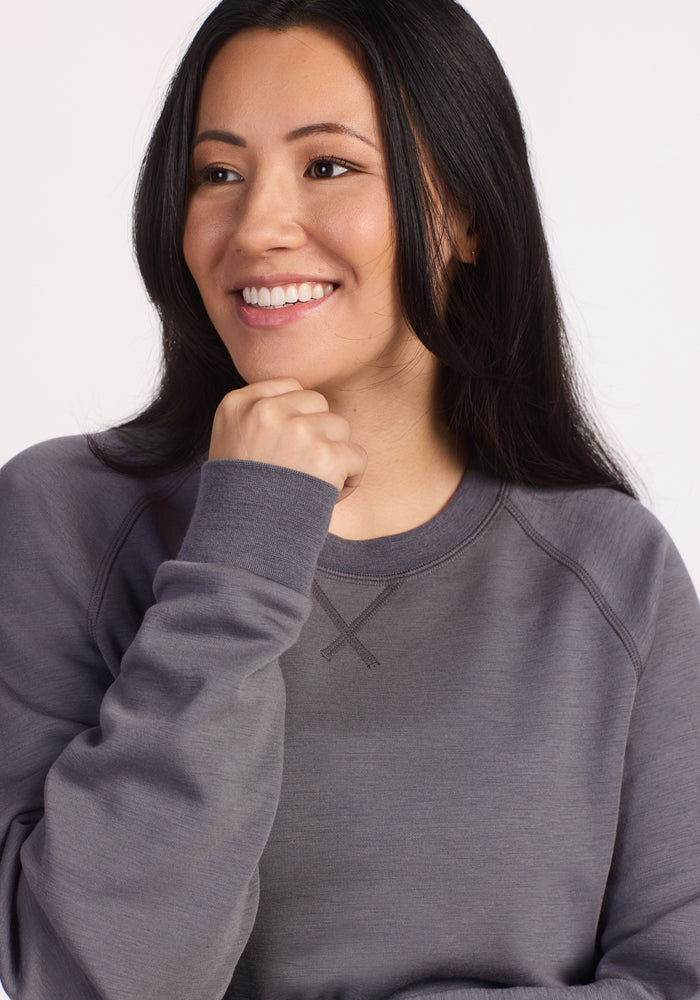A woman with long dark hair smiles and looks to the side, her chin resting on her hand. She wears the Woolx Bailey Crewneck Sweatshirt in Arctic Dusk, featuring a loose fit and long sleeves against a plain light background.
