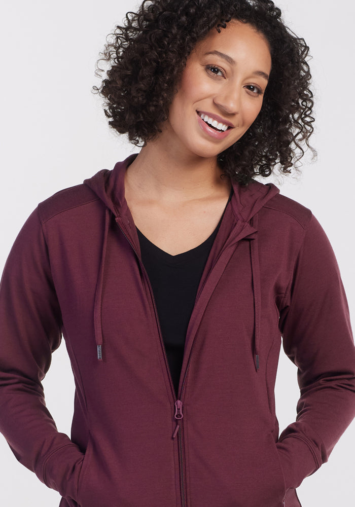 A woman with curly hair smiles while wearing the Woolx Ryann Hoodie in Grape Harvest over a black top, standing against a plain white background.