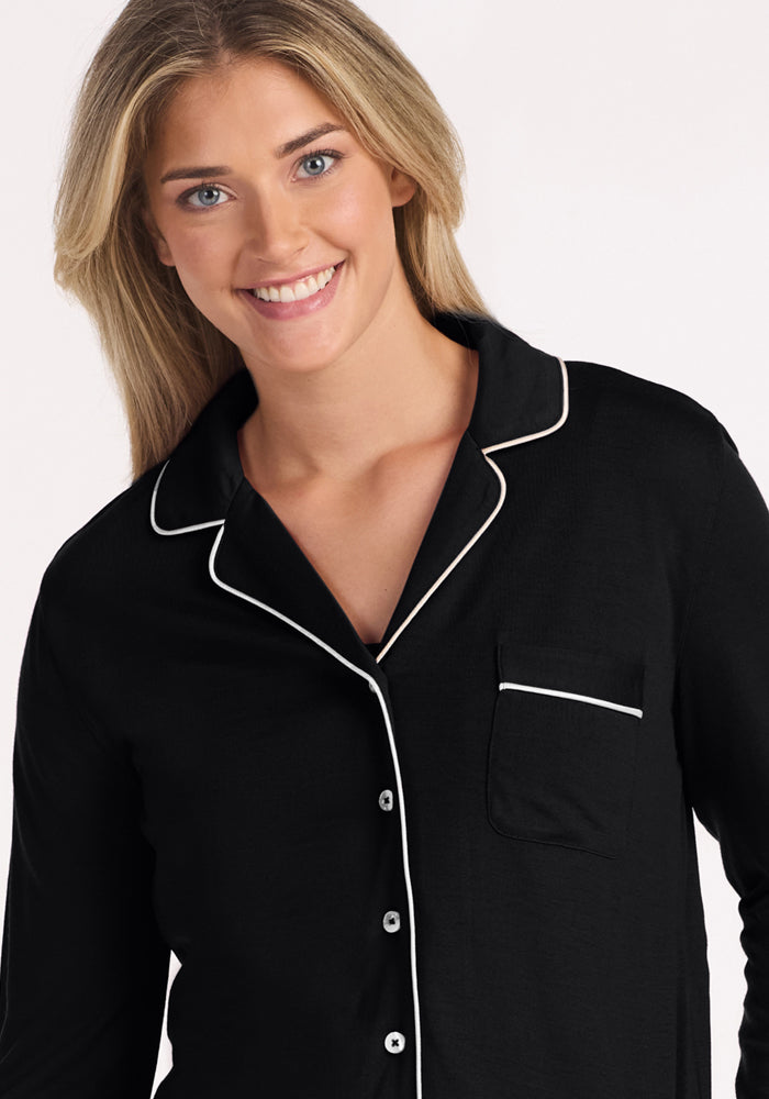 A smiling person with long blonde hair wears the Woolx Harper FeatherTouch® Pajama Set in black with white piping and buttons, standing against a plain, light background.