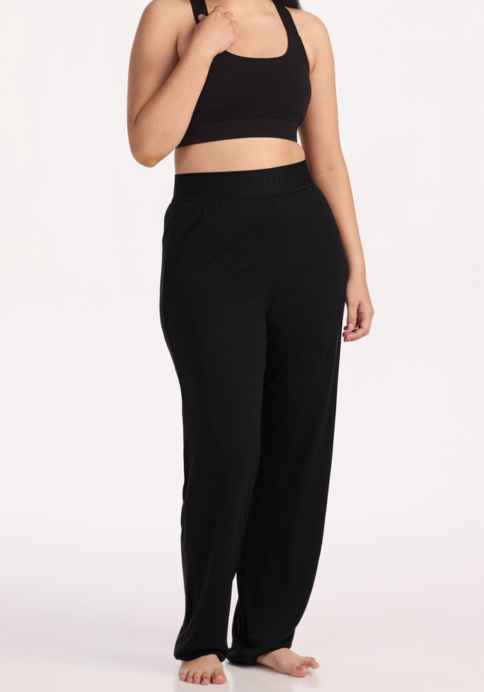 A person stands barefoot against a plain white background wearing Woolx Maya Lounge Pants in black, paired with a black sports bra. The image shows their body from shoulders to feet.