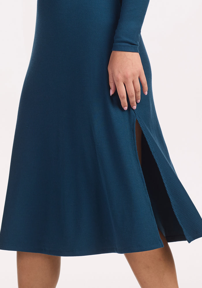 A person wears the Woolx Caroline Ribbed Twirl Dress in Majolica Blue, featuring a side slit and temperature-regulating merino fabric. Their hand rests near the hip, standing out against a plain white background.